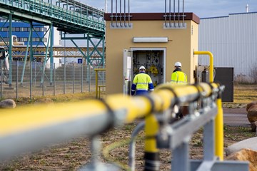 Wasserstoffprojekt HYPOS: H2-Netz im Vordergrund befinden sich Rohrleitungen, im Hintergrund Männer mit Arbeitskleidung in einer technischen Anlage