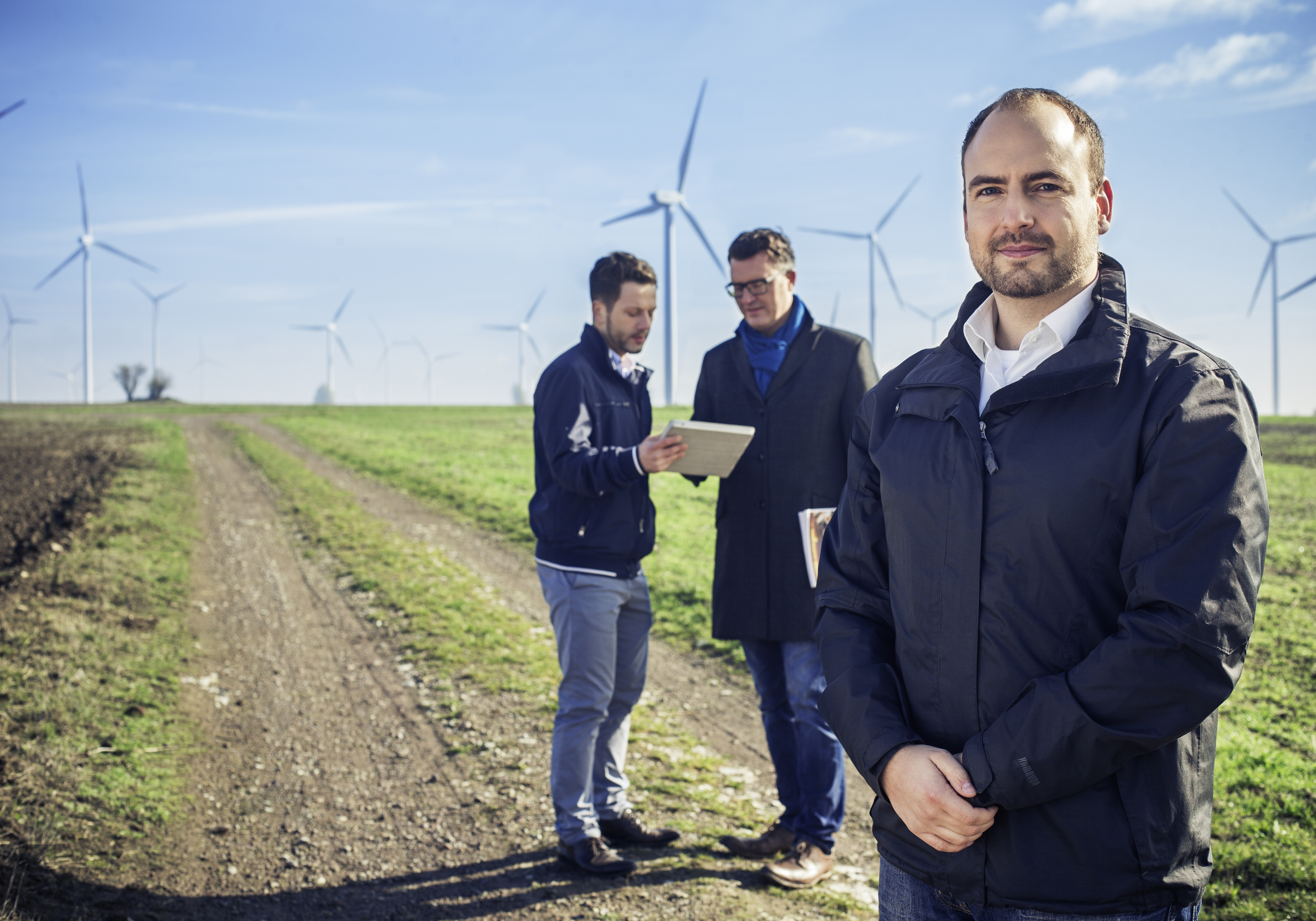 Frank Eberlein von envia THERM steht mit 2 weiteren Männern auf einem Feldweg vor zahlreichen Windkrafträdern