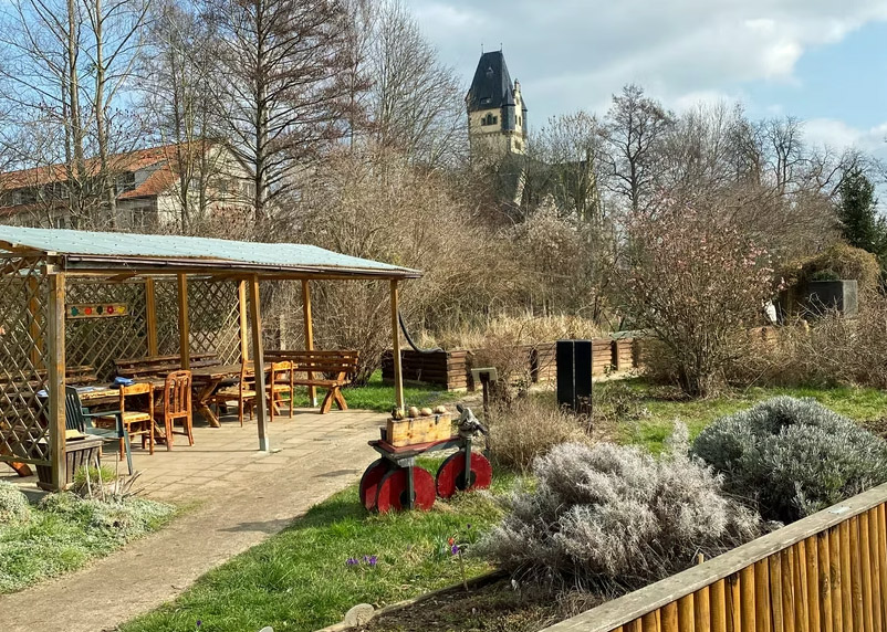 Blick in den  Ökogarten Quedlinburg, im Hintergrund eine Sitzgruppe mit Stühlen und Tischen, im Vordergrund Pflanzenstauden