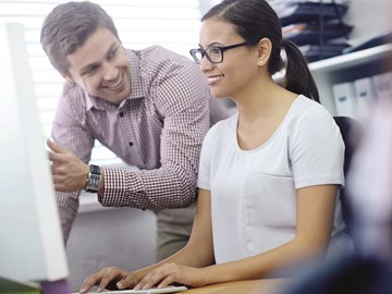 Frau sitzt vor Monitor, Mann steht daneben Frau sitzt vor Monitor, Mann steht daneben