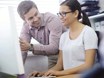 Frau sitzt vor Monitor, Mann steht daneben Frau sitzt vor Monitor, Mann steht daneben